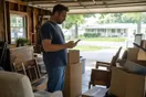 A man stands in his garage on a Saturday morning surrounded by boxes and old furniture, looking at his phone to find someone to haul it away today.