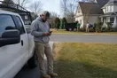 A landscaper stands beside his parked truck in a suburban driveway checking his phone while a rival crew works a yard down the street, in a natural documentary style.