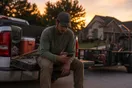 A landscaper stands in his truck bed at dusk scrolling a quiet phone after a long install day, in a natural documentary style.
