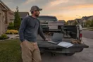 A landscaping company owner standing beside his truck at dusk, scrolling a laptop with a stack of unsent estimates on the tailgate.