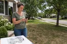 A homeowner stands in their front yard holding a phone, comparing landscaping company websites in natural daylight, documentary style.