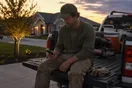 A landscaper sits on his truck's tailgate scrolling a slow-loading website on his phone after a long install day, in a natural documentary style.