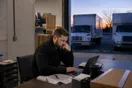 A moving company owner checks an empty booking calendar in a warehouse office next to parked trucks at dusk, in a natural documentary style.