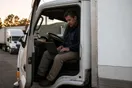 A moving company owner sitting in the cab of a parked truck at dusk, phone in hand, laptop balanced on the dashboard beside a stack of paper quotes.