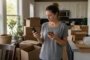 A person stands in a half-packed kitchen surrounded by boxes, holding a phone while comparing moving company websites, photographed in a natural documentary style.