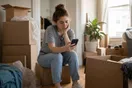A person sits on a half-packed cardboard box in a living room, phone in hand, waiting for a moving company's website to load.