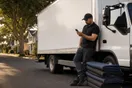 A moving company owner leans against a parked box truck checking a slow-loading page on his phone while a stack of moving blankets sits nearby, in a natural documentary style.