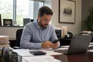 An accountant checks his phone at his desk in a small office during tax season, papers and a laptop nearby, natural documentary style.
