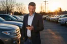 A used car dealership owner checks his phone while standing on his lot at sunrise among rows of vehicles, natural documentary style.