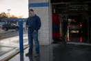 A car wash owner checks his phone near the tunnel entrance of his wash location in early morning light, natural documentary style.