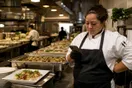 A caterer checks her phone beside trays of plated food in a commercial kitchen before a delivery, natural documentary style.