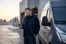 A courier company owner checks his phone beside a parked delivery van outside a warehouse dock early in the morning, natural documentary style.