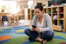 A daycare director sits on a classroom rug at closing time reviewing her phone, small chairs and cubbies visible behind her, natural documentary style.
