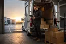 A delivery company owner stands at the back of an open cargo van in a warehouse loading bay at dawn, checking a tablet, natural documentary style.
