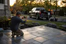 A landscaper kneels beside a finished stone patio at golden hour, phone in hand photographing the work, truck and tools visible in the driveway behind him, natural documentary style.