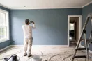 A painter steps back from a freshly painted living room wall to check the color under afternoon light, phone in hand snapping a photo, drop cloths and ladder visible, natural documentary style.