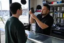 A repair shop owner stands at his counter holding a cracked phone up to the light, a customer waiting across the glass, morning light through the storefront window, natural documentary style.