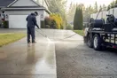 A pressure washer stands on a driveway holding a wand mid-spray on a half-cleaned concrete surface, the clean and dirty halves visible in morning light, trailer and hose reel nearby, natural documentary style.