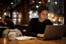 A startup founder checking an accountant's website on a laptop at a coffee shop table, notebook and coffee nearby, natural documentary style.