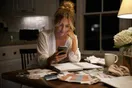 A bride-to-be scrolling a bakery's website on her phone at a kitchen table covered in wedding planning notes, natural documentary style.