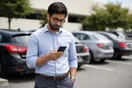 A man in a driveway checking a car dealership website on his phone before visiting the lot his coworker recommended, natural documentary style.