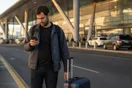A traveler checking a car rental company's website on his phone at an airport curb after a friend recommended them, natural documentary style.