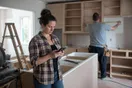 A homeowner standing in her unfinished kitchen looking up a contractor's website on her phone while a builder measures cabinets in the background, natural documentary style.