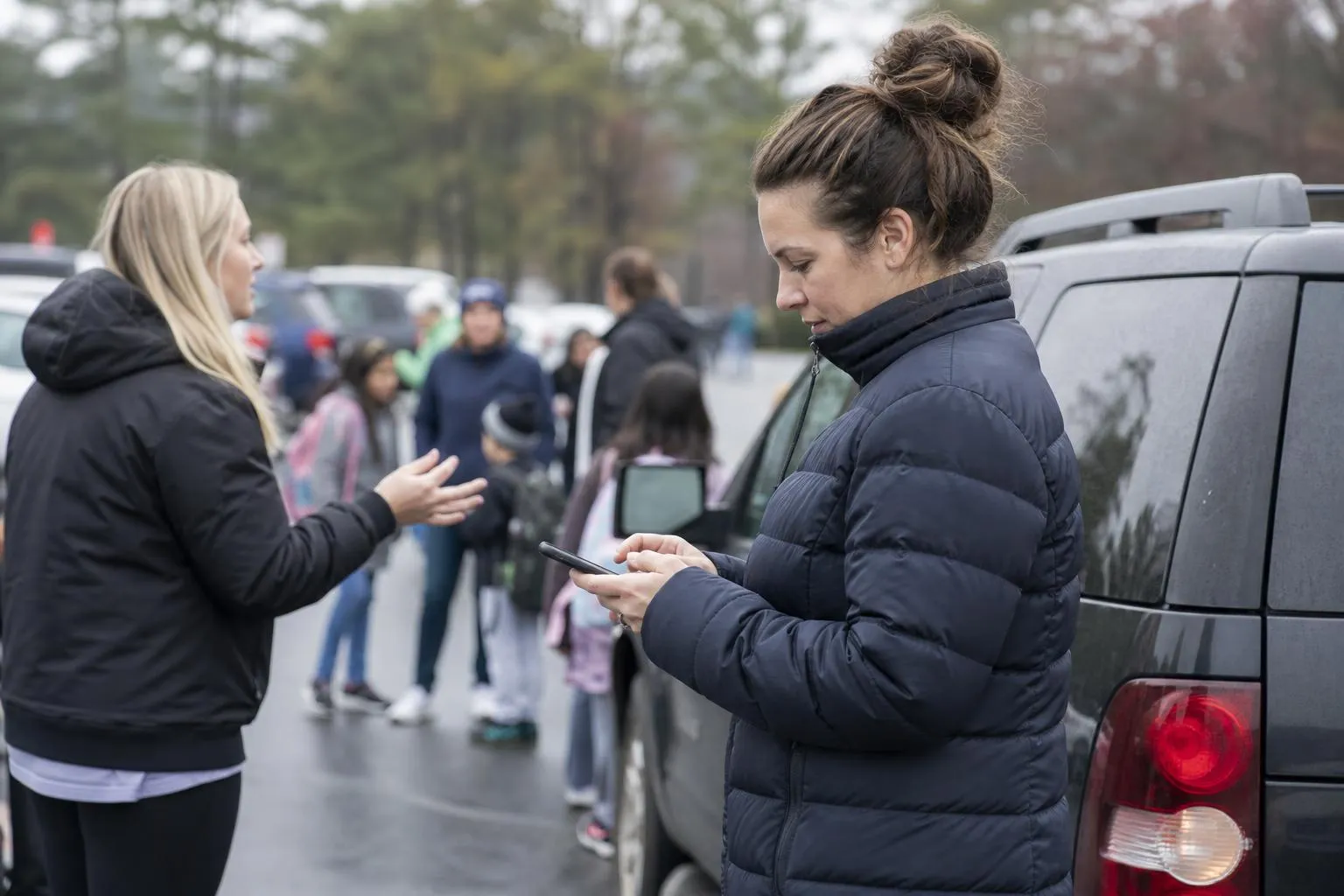 A mother standing at a school pickup line scrolling her phone while another parent gestures, both bundled up on a cool morning.