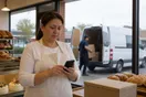 A bakery owner in a Chicago strip mall scrolling a delivery company's website on her phone while a driver loads a van in the background, natural documentary style.