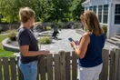 A homeowner standing in her backyard pointing out a neighbor's new patio while checking a landscaping company's name on her phone, natural documentary style suburban afternoon.