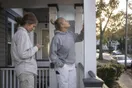 A homeowner and her neighbor standing on a porch admiring freshly painted exterior trim while one looks up a painting company on her phone, natural documentary style.