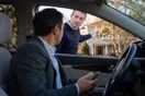 A woman on her phone in a parked car outside a house with a friend gesturing nearby, natural documentary style American suburb.
