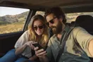 A couple checking a winery's website on a phone in the passenger seat of a car driving through vineyard hills, natural documentary style.