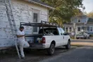 A painter stands beside his ladder and parked truck outside a house checking his phone while a competitor's crew works a repaint down the block, in a natural documentary style.