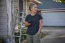 A homeowner stands beside a ladder in a driveway holding a phone, comparing painting contractor websites in daylight, documentary style.