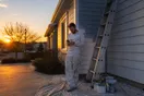 A painter stands beside a ladder outside a freshly painted house, checking messages on his phone as the sun sets.