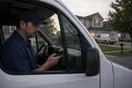 A pest control technician sits in his parked service truck outside a house checking his phone while a competitor's van pulls into a driveway across the street, in a natural documentary style.
