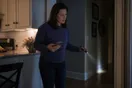 A homeowner stands in a kitchen doorway looking at their phone with a worried expression, a flashlight in the other hand pointed toward a baseboard, in dim evening light.