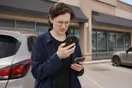 A person stands outside a strip mall storefront squinting at a cracked phone screen while typing on another phone in their other hand, in a natural documentary style.