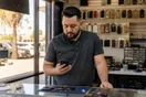 A phone repair shop owner stands behind the counter checking a quiet phone late in the afternoon, in a natural documentary style.