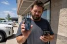 A person squints at a spiderwebbed phone screen while holding a second phone, photographed in a natural documentary style, no readable text visible.