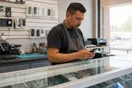 A repair shop owner stands behind his counter checking his phone near a display case of devices during a slow afternoon.
