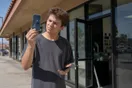 A young customer holds a cracked phone up to the light outside a small repair shop storefront, glancing at their other phone's screen, in natural afternoon light.