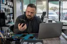 A repair shop owner squints at a cracked phone screen mirrored against a slow-loading laptop on the counter, in a natural documentary style.