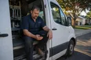 A plumber sits in his parked work van between jobs, checking his phone on a quiet suburban street in late afternoon light.