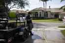 A pressure washing operator stands beside his parked trailer rig on a driveway checking his phone while a competitor works a roof across the street, in a natural documentary style.