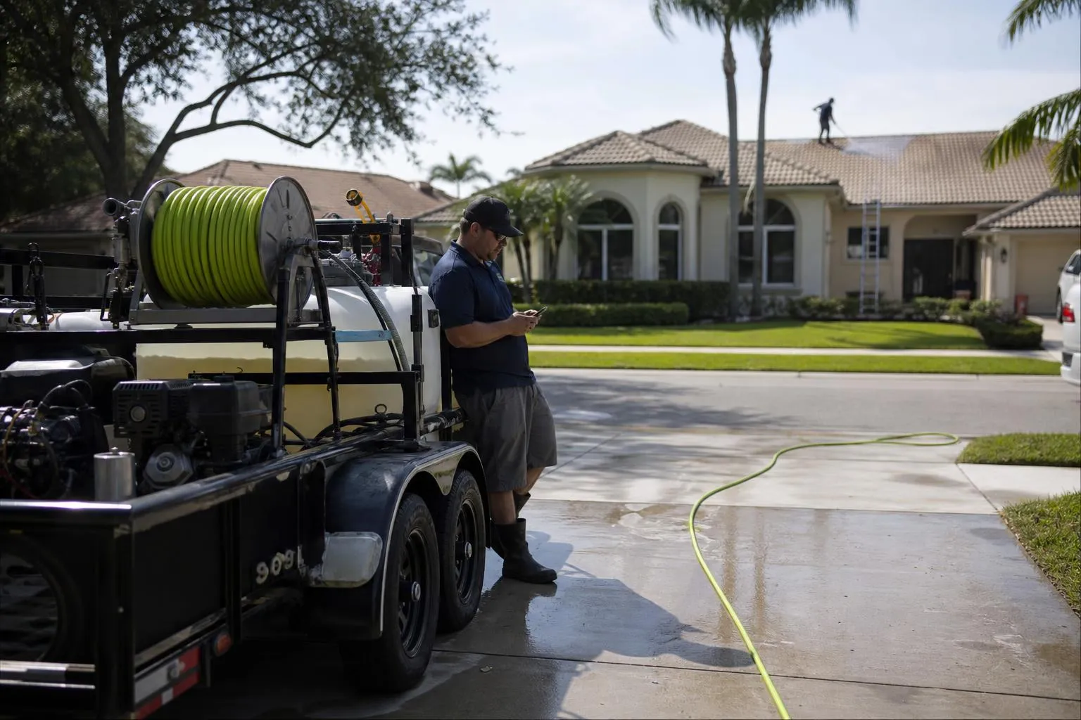 A pressure washing operator stands beside his parked trailer rig on a driveway checking his phone while a competitor works a roof across the street, in a natural documentary style.