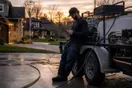 A pressure washing business owner leans against his trailer at dusk checking a quiet phone after a spring route, in a natural documentary style.