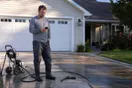 A person stands on a driveway looking at a phone with a pressure washer parked nearby, natural daylight, documentary style.