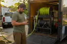 A pressure washing contractor loads hoses into his trailer in a driveway at golden hour, checking his phone before the next stop.